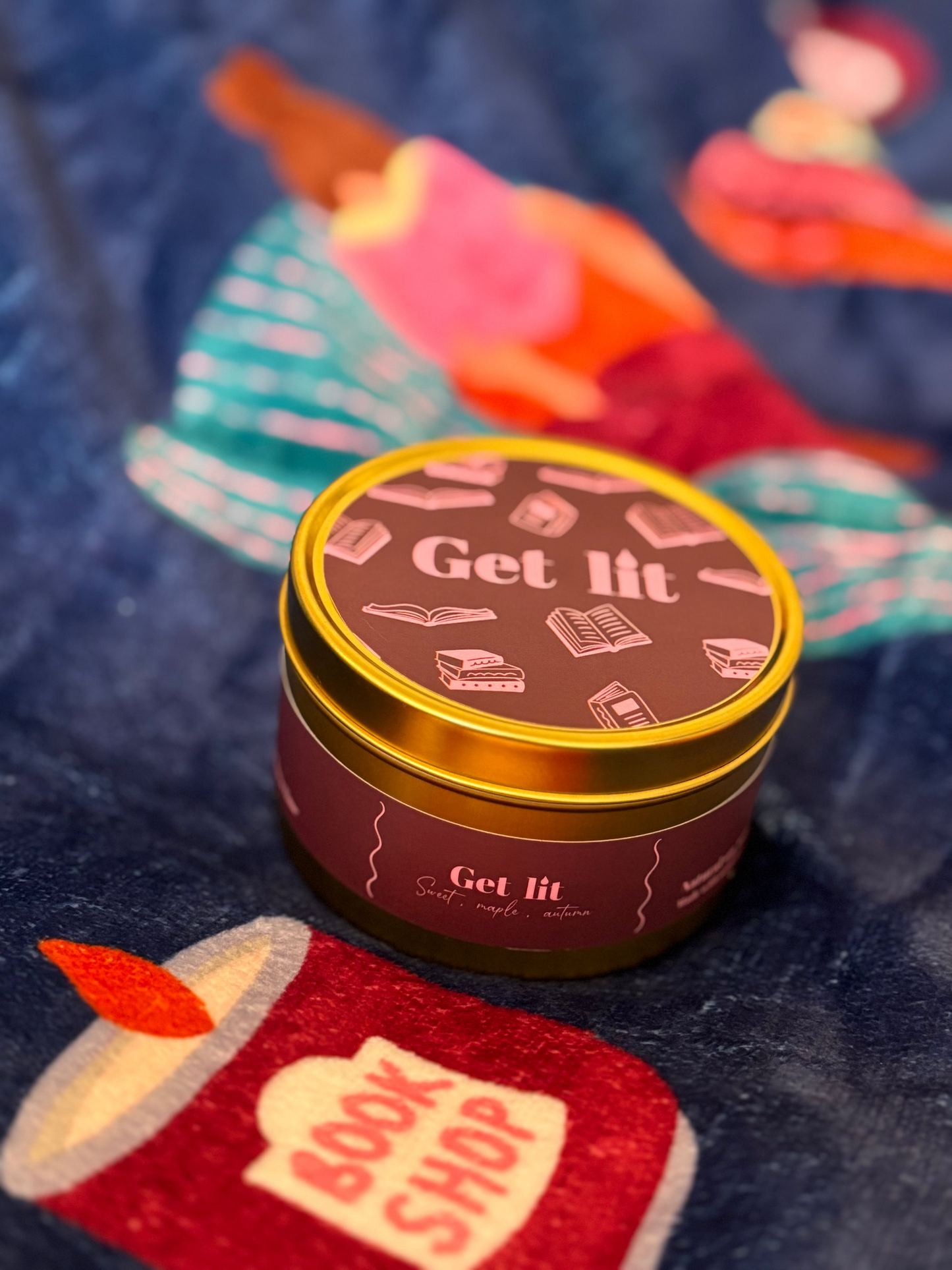 Candle tin labeled 'Get Lit' on a dark fabric background with colorful items.